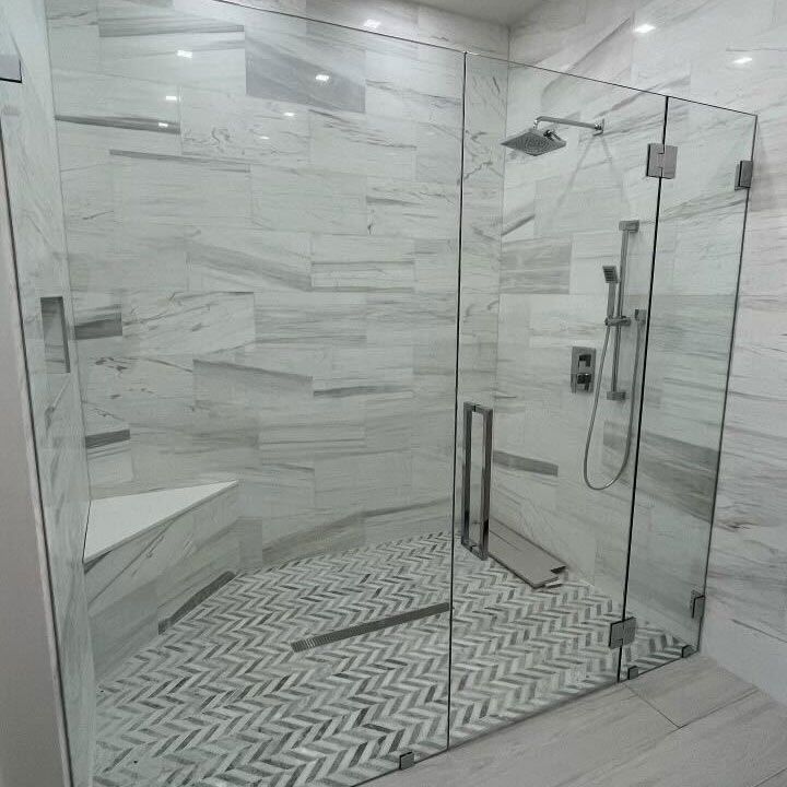 Frameless glass shower enclosure with polished chrome hardware, a built-in corner bench and a herringbone marble mosaic floor